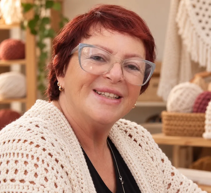 Marlete Souza sorrindo no ateliê, com novelos nas prateleiras ao fundo